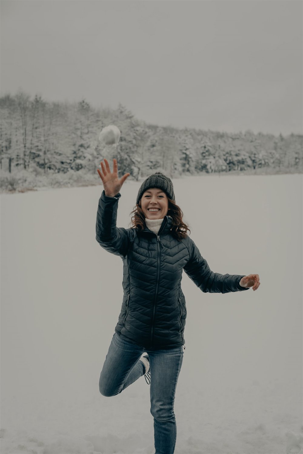 Smilende kvinne  med lue, jakke og jeans kaster snøball mot fotografen